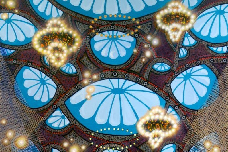 A detail of Michiko Itatani's "'Bluelift' painting from Cosmic Theater 23-B-5," 2023, an oil on canvas work of a liminal space that looks like an expansive lobby with sacred geometry and many lit chandeliers hanging from the ceiling.